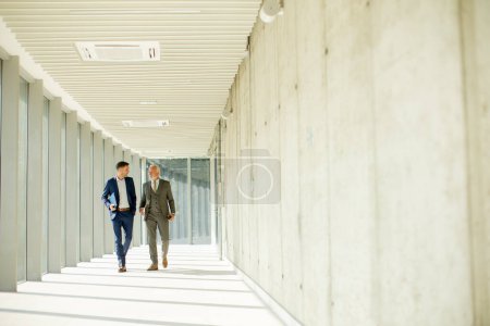 Photo for Young and a senior businessman walk down an office hallway, deep in conversation. They are both dressed professionally, reflecting their business acumen and status. They are navigating the complexities of the corporate world, finding solutions and ma - Royalty Free Image