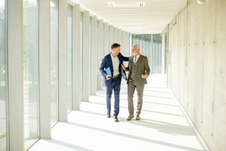 Photo for Young and a senior businessman walk down an office hallway, deep in conversation. They are both dressed professionally, reflecting their business acumen and status. They are navigating the complexities of the corporate world, finding solutions and ma - Royalty Free Image