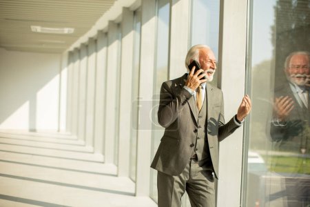 Foto de A senior business man stands in an office hallway, focused on his mobile phone. He is dressed in formal attire, exuding confidence and professionalism - Imagen libre de derechos
