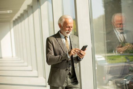 Téléchargez les photos : A senior business man stands in an office hallway, focused on his mobile phone. He is dressed in formal attire, exuding confidence and professionalism - en image libre de droit