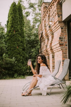 Téléchargez les photos : Pretty young woman with curly hair relaxes on a deck chair and enjoys a glass of fresh lemonade - en image libre de droit