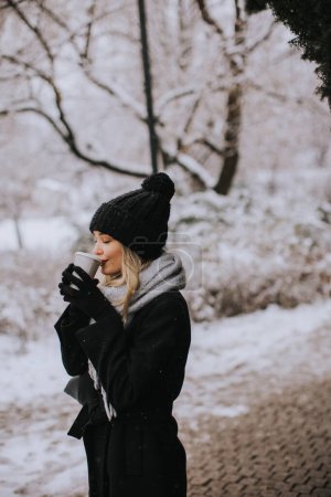 Foto de Una mujer joven se encuentra en un país de las maravillas de invierno, vistiendo ropa de abrigo y tomando café caliente para ir - Imagen libre de derechos