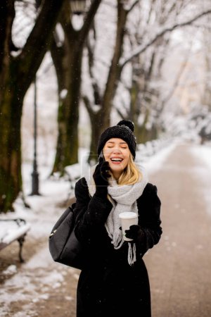 Téléchargez les photos : Une jeune femme portant des vêtements chauds d'hiver et un chapeau tricoté sourit joyeusement alors qu'elle se tient dans la neige et utilise un téléphone portable tout en tenant une tasse de cofee - en image libre de droit
