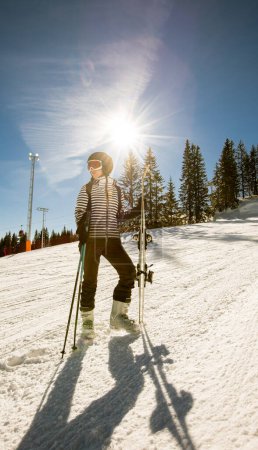 Foto de Una joven soltera disfruta de un soleado día de invierno de esquí, vestida con equipo de nieve completo con botas de esquí y gafas de sol. - Imagen libre de derechos