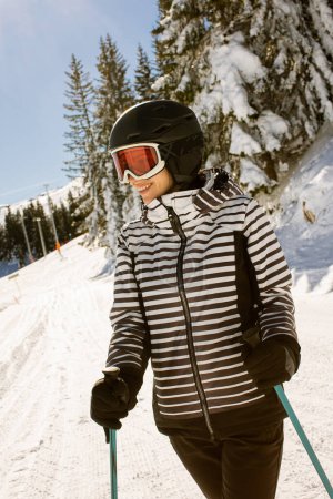 Foto de Joven mujer disfrutando de un día de invierno esquiando en las pistas cubiertas de nieve, rodeada de árboles altos y vestida para temperaturas frías - Imagen libre de derechos