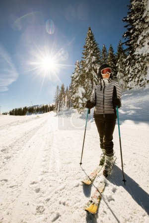 Foto de Una joven soltera disfruta de un soleado día de invierno de esquí, vestida con equipo de nieve completo con botas de esquí y gafas de sol - Imagen libre de derechos