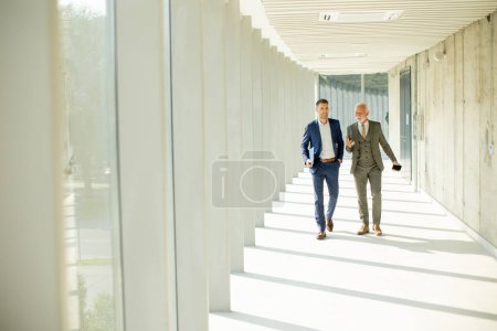 Foto de Young y un hombre de negocios mayor caminan por el pasillo de una oficina, en lo profundo de la conversación. Ambos están vestidos profesionalmente, reflejando su perspicacia y estatus empresarial. Están navegando por las complejidades del mundo corporativo, encontrando soluciones y - Imagen libre de derechos
