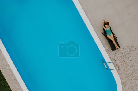 Téléchargez les photos : Vue aérienne sur une jolie jeune femme allongée sur un pont d'ombrage près de la piscine dans la cour arrière de la maison - en image libre de droit