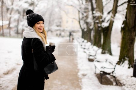 Téléchargez les photos : A young woman stands in a winter wonderland, wearing warm clothing and drinking hot coffee to go - en image libre de droit
