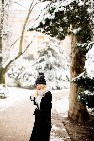 Photo for A young woman stands in a winter wonderland, wearing warm clothing and drinking hot coffee to go - Royalty Free Image