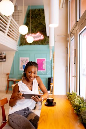 Téléchargez les photos : Jolie jeune femme noire buvant du café tout en regardant tablette numérique dans le café - en image libre de droit