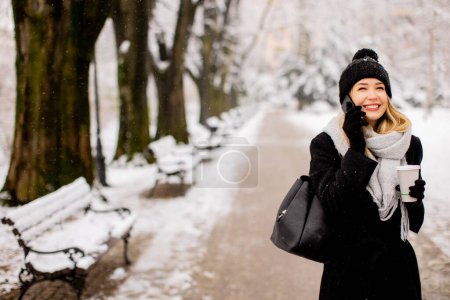 Photo for A young woman wearing warm winter clothes and a knit hat smiles happily as she stands in the snow and using mobile phone whil holding cofee cup - Royalty Free Image
