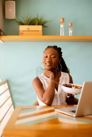 Foto de Mujer negra bastante joven desayunando saludable mientras trabaja en el ordenador portátil en la cafetería - Imagen libre de derechos