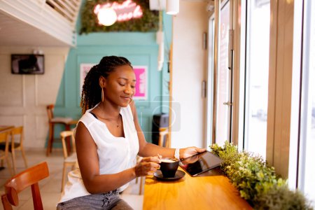 Foto de Mujer negra bastante joven bebiendo café mientras mira la tableta digital en el café - Imagen libre de derechos