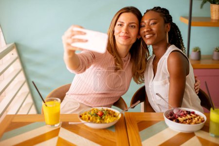 Photo for Two pretty young women, caucasian and black one, taking selfie with mobile phone in the cafe - Royalty Free Image