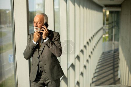 Foto de A senior business man stands in an office hallway, focused on his mobile phone. He is dressed in formal attire, exuding confidence and professionalism - Imagen libre de derechos