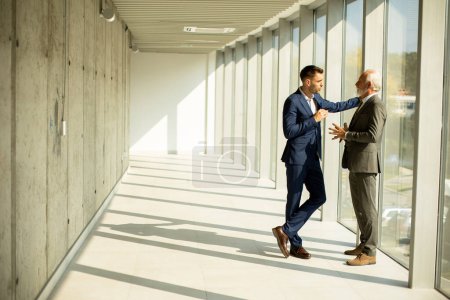 Téléchargez les photos : Beau homme d'affaires mature et son jeune collègue discutant dans le couloir du bureau - en image libre de droit