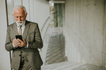 Téléchargez les photos : A senior business man stands in an office hallway, focused on his mobile phone. He is dressed in formal attire, exuding confidence and professionalism - en image libre de droit