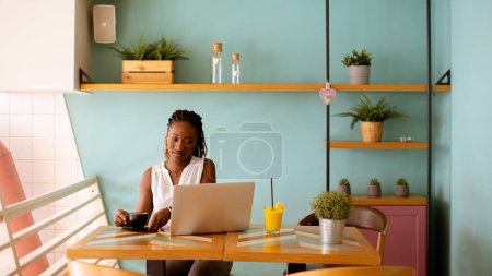 Téléchargez les photos : Jolie jeune femme noire buvant du café tout en travaillant sur un ordinateur portable dans le café - en image libre de droit