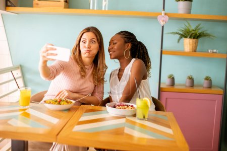 Photo for Two pretty young women, caucasian and black one, taking selfie with mobile phone in the cafe - Royalty Free Image