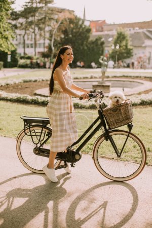 Photo for Pretty young woman with white bichon frise dog in the basket of electric bike - Royalty Free Image