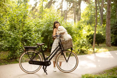 Photo for Pretty young woman putting white bichon frise dog in the basket of electric bike - Royalty Free Image