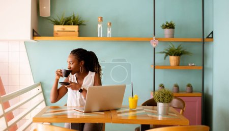 Foto de Mujer negra bastante joven bebiendo café mientras trabaja en el portátil en el café - Imagen libre de derechos