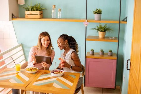 Téléchargez les photos : Deux jolies jeunes femmes noires et caucasiennes s'amusent, boivent des jus de fruits frais et prennent un petit déjeuner sain au café - en image libre de droit