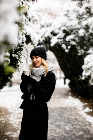 Téléchargez les photos : Jolie jeune femme en vêtements chauds jouissant dans la neige avec tasse à café à emporter - en image libre de droit