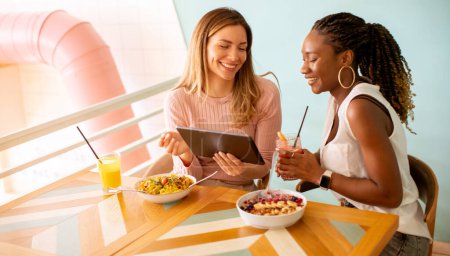 Téléchargez les photos : Deux jolies jeunes femmes noires et caucasiennes s'amusent, boivent des jus de fruits frais et prennent un petit déjeuner sain au café - en image libre de droit
