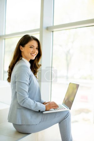 Pretty young business woman sitting on the stairs on office hallway and working on laptop