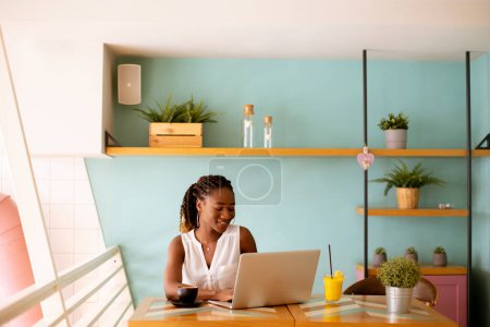 Téléchargez les photos : Jolie jeune femme noire buvant du café tout en travaillant sur un ordinateur portable dans le café - en image libre de droit