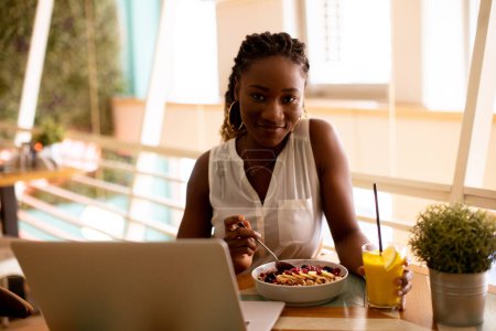 Téléchargez les photos : Jolie jeune femme noire prenant un petit déjeuner sain tout en travaillant sur ordinateur portable dans le café - en image libre de droit