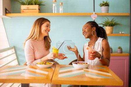 Foto de Dos mujeres negras y caucásicas bastante jóvenes pasándola bien, bebiendo jugos frescos y desayunando sano en la cafetería - Imagen libre de derechos