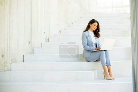 Téléchargez les photos : Jolie jeune femme d'affaires assise dans les escaliers sur le couloir du bureau et travaillant sur un ordinateur portable - en image libre de droit