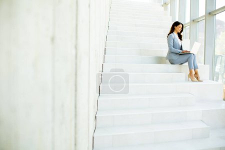 Foto de Mujer de negocios bastante joven sentado en las escaleras en el pasillo de la oficina y trabajando en el ordenador portátil - Imagen libre de derechos