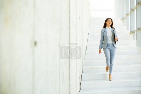 Téléchargez les photos : Jeune femme d'affaires marchant dans les escaliers et tenant un ordinateur portable dans le couloir du bureau - en image libre de droit