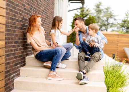 Téléchargez les photos : Famille avec une mère, un père, un fils et une fille assis dehors sur les marches d'un porche d'une maison en brique - en image libre de droit
