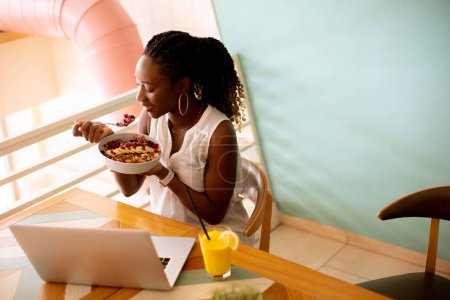 Foto de Mujer negra bastante joven desayunando saludable mientras trabaja en el ordenador portátil en la cafetería - Imagen libre de derechos