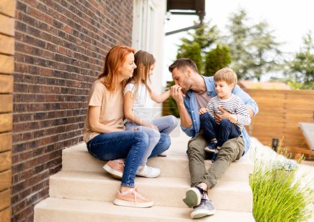 Foto de Familia con una madre, padre, hijo e hija sentados fuera en las escaleras de un porche delantero de una casa de ladrillo - Imagen libre de derechos