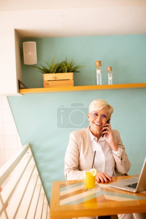 Photo for Senior woman using mobile phone while working on the laptop and drinking fresh orange juice in the cafe - Royalty Free Image