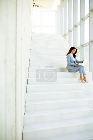 Photo for Pretty young business woman sitting on the stairs on office hallway and working on laptop - Royalty Free Image
