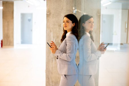 Téléchargez les photos : Jolie jeune femme d'affaires utilisant un téléphone portable dans le couloir du bureau - en image libre de droit