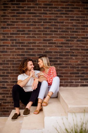 Téléchargez les photos : Beau jeune couple souriant amoureux assis devant le mur de briques de la maison - en image libre de droit