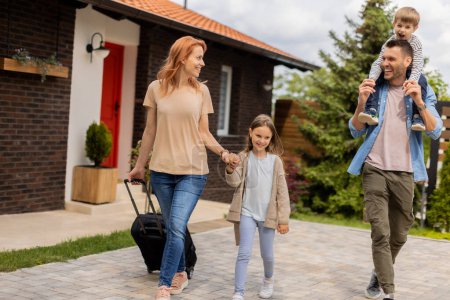 Téléchargez les photos : Famille avec une mère, père, fils et fille marchant avec des bagages à l'extérieur sur le porche d'une maison en brique - en image libre de droit