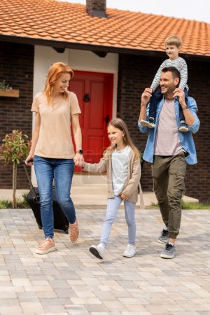 Photo for Family with a mother, father, son and daughter walking with  abaggage outside on the front porch of a brick house - Royalty Free Image