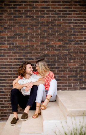 Téléchargez les photos : Beau jeune couple souriant amoureux assis devant le mur de briques de la maison - en image libre de droit