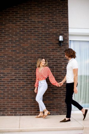 Téléchargez les photos : Beau jeune couple souriant amoureux marchant devant le mur de briques de la maison - en image libre de droit
