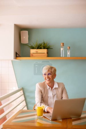 Photo for Senior woman using mobile phone while working on the laptop and drinking fresh orange juice in the cafe - Royalty Free Image