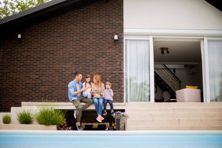 Photo for Family with a mother, father, son and daughter sitting outside by the swimming pool and eating strawberries - Royalty Free Image